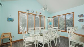 A dining area with a round glass table and white chairs, large windows with blinds, and a ceiling fan.