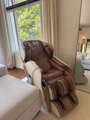 A massage chair in a modern living room with a large window, a sofa, and a mirror.