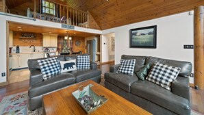 A spacious living area with a high ceiling and wooden beams, featuring two leather sofas with checkered pillows, a wooden coffee table, and a kitchen area with wooden cabinets and a visible staircase.