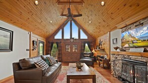 A spacious living room with a high ceiling, wooden beams, a fireplace, and large windows.