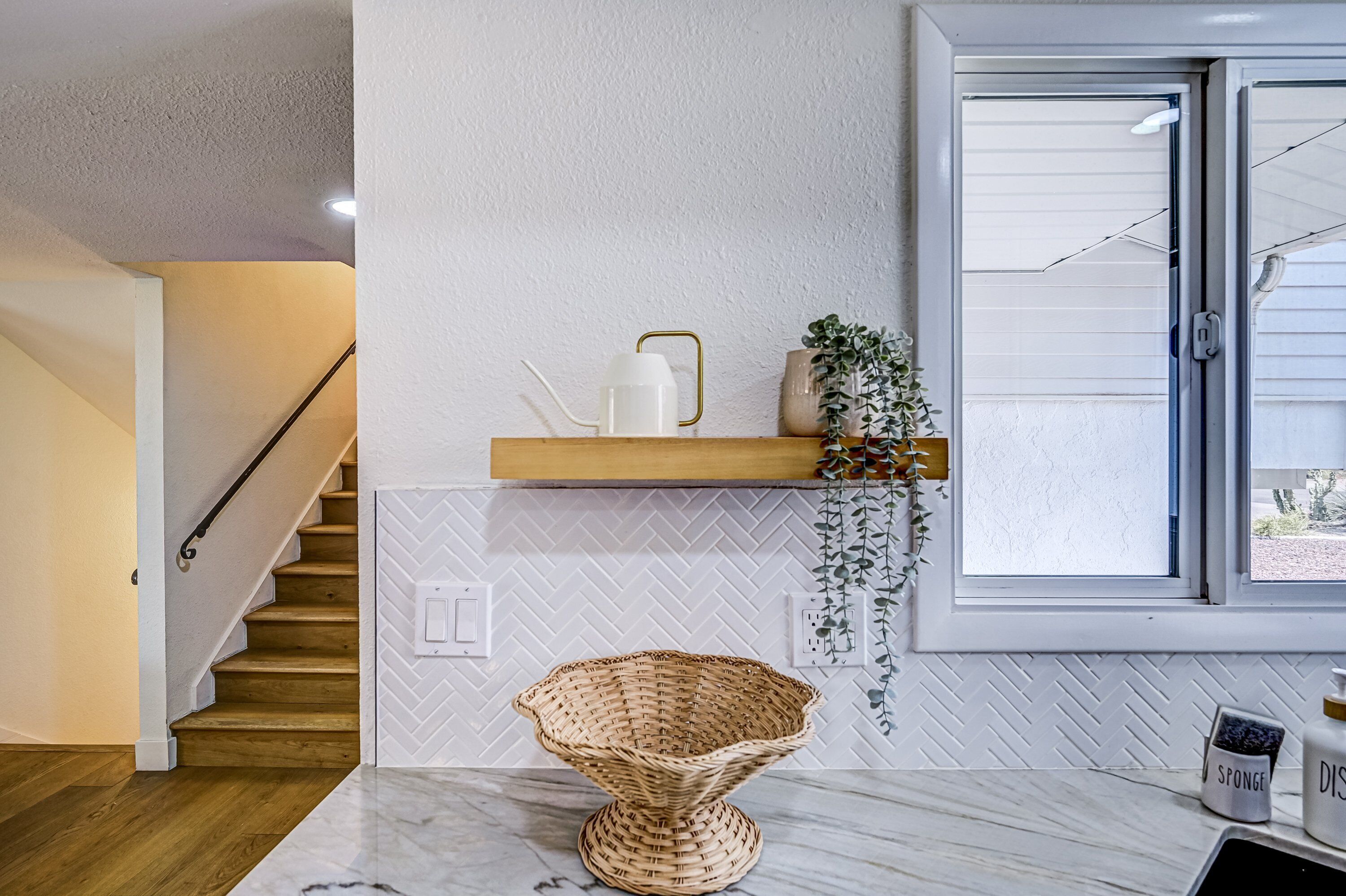 A kitchen with a herringbone-patterned backsplash, a wooden staircase, and a window with a view.