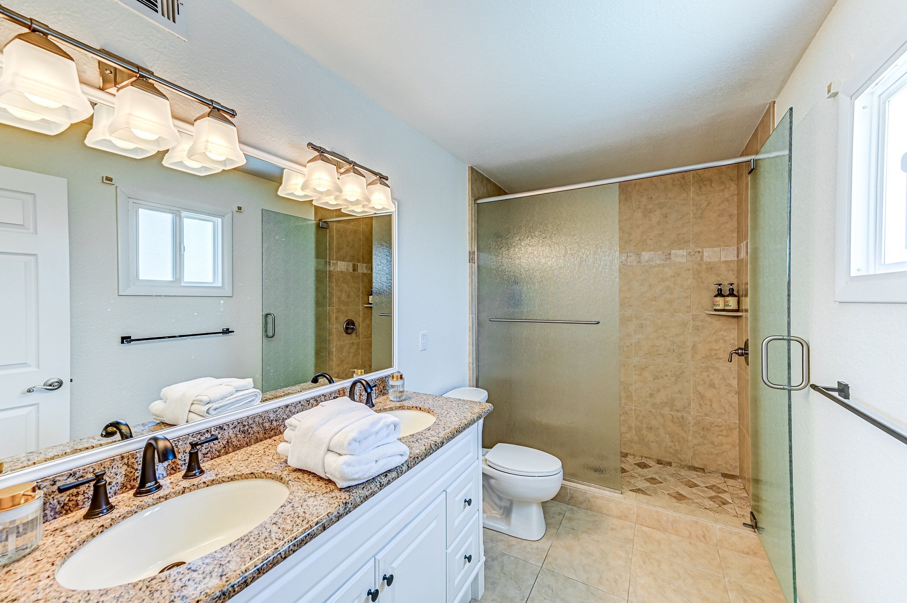 A modern bathroom with a double sink vanity, a large mirror, a glass-enclosed shower, and a toilet.