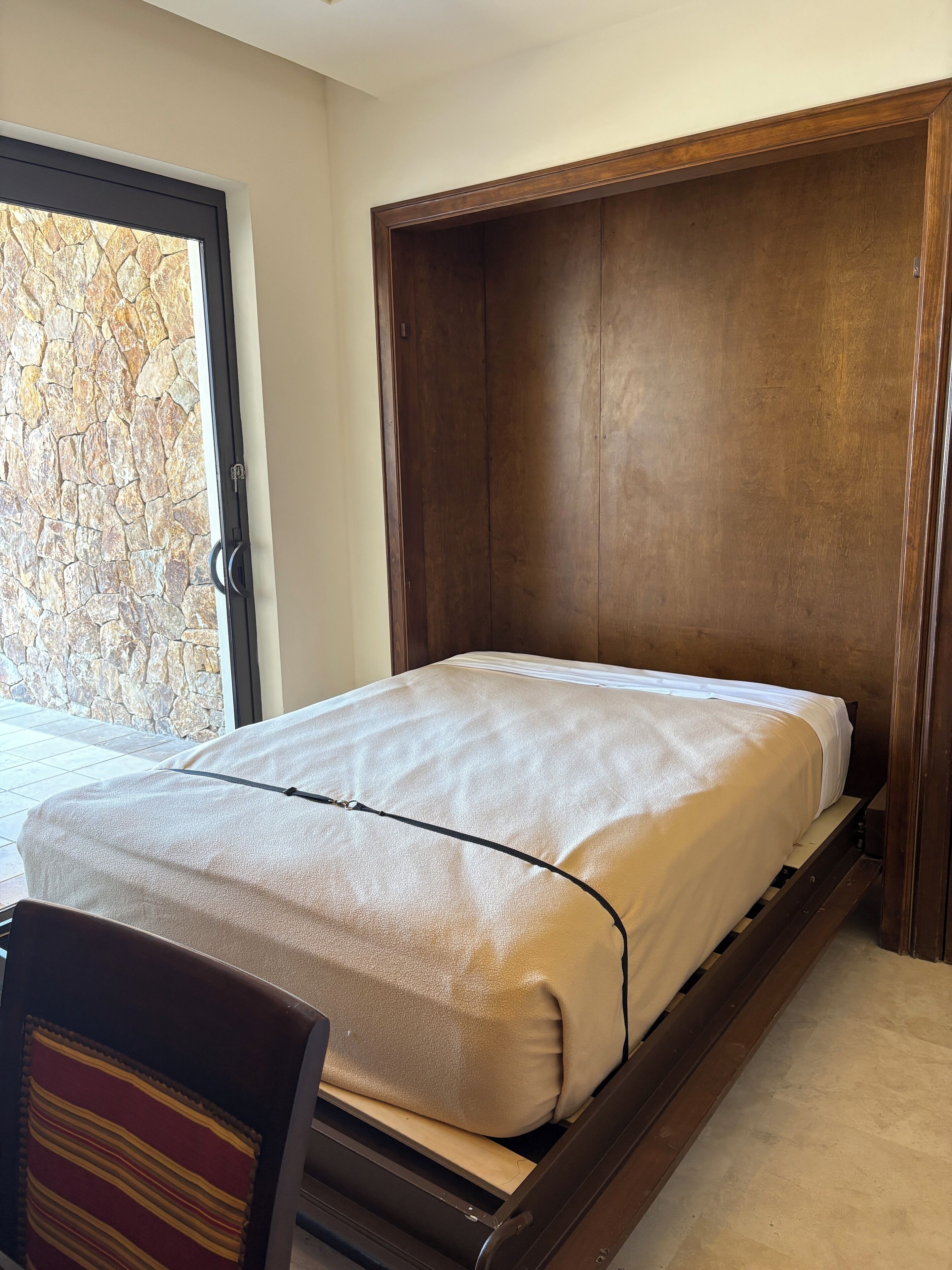 A bedroom with a wooden bed frame, a stone wall, and a view through a glass door.