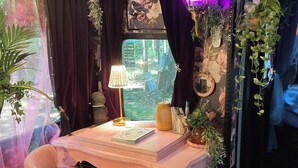 Una habitación acogedora con un escritorio rosa, una silla cómoda, una ventana con cortinas y varias plantas y adornos.