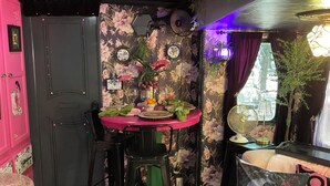 Una habitación compacta con una mesa de comedor dispuesta para dos personas, un aparador rosa, papel tapiz floral y una ventana con cortinas.
