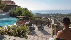A poolside area with a wooden deck, lounge chairs, and a view of the sea.