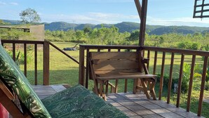 A wooden deck with a bench and a lounge chair, overlooking a grassy area and distant hills.