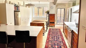 Une cuisine moderne avec un îlot central, un coin repas avec des chaises, un tapis à motifs et une vue sur l’extérieur par une fenêtre.