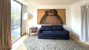 Un salon moderne avec un canapé bleu foncé, un tapis à motifs, une grande peinture abstraite, un lampadaire et une vue dégagée sur l’extérieur grâce à une grande fenêtre.
