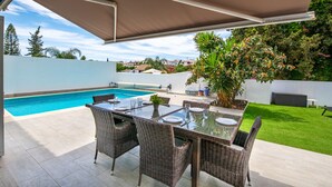 Un patio exterior con piscina, mobiliario de mimbre y una zona cubierta.