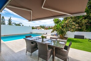 Un patio exterior con piscina, mobiliario de mimbre y una zona cubierta.