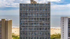 Un hôtel à plusieurs étages doté d’une façade moderne en verre, situé près d’une plage, avec un parking visible et une vue dégagée sur l’océan.