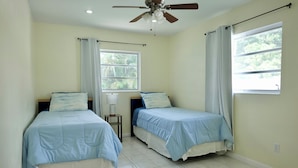 Una habitación con dos camas, ventilador de techo, ventana con cortinas y mesita de noche con lámpara.