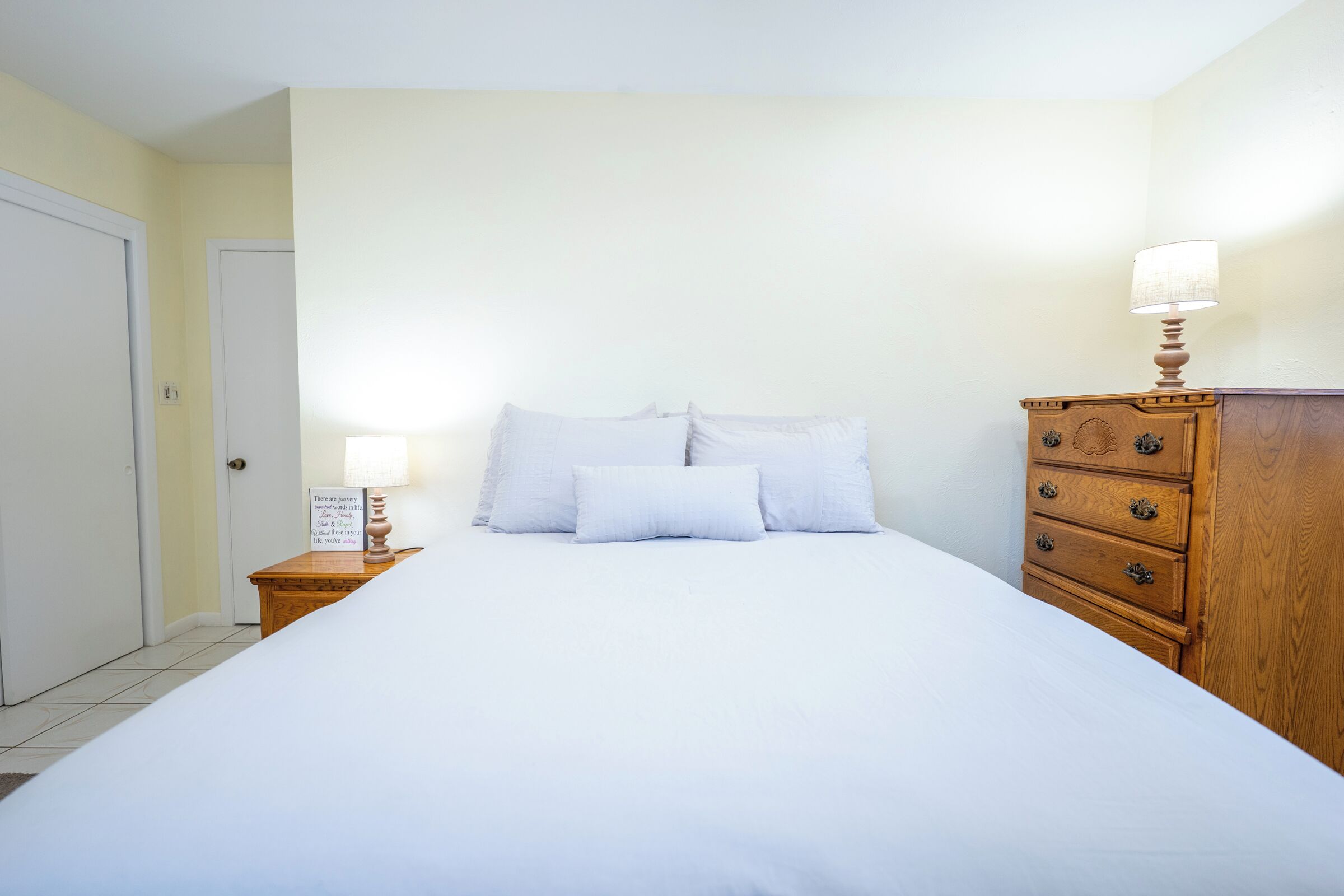 A neatly made bed with white linens, a wooden dresser with drawers, and a bedside lamp on a nightstand.