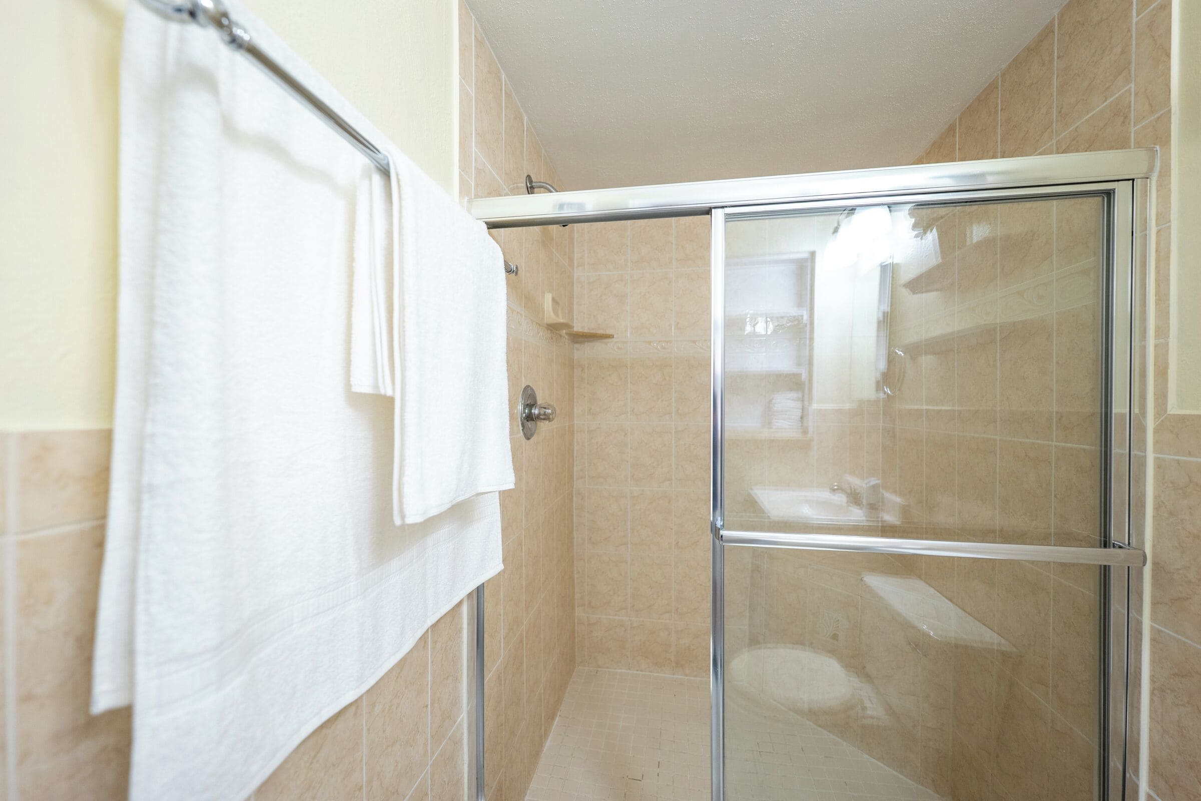 Cabina de ducha con paredes de vidrio, azulejos color beige, una toalla blanca en un perchero y cabezal de ducha visible.