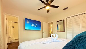 Une chambre d’hôtel avec un lit, un ventilateur de plafond, un téléviseur diffusant Disney+ et un miroir au mur.