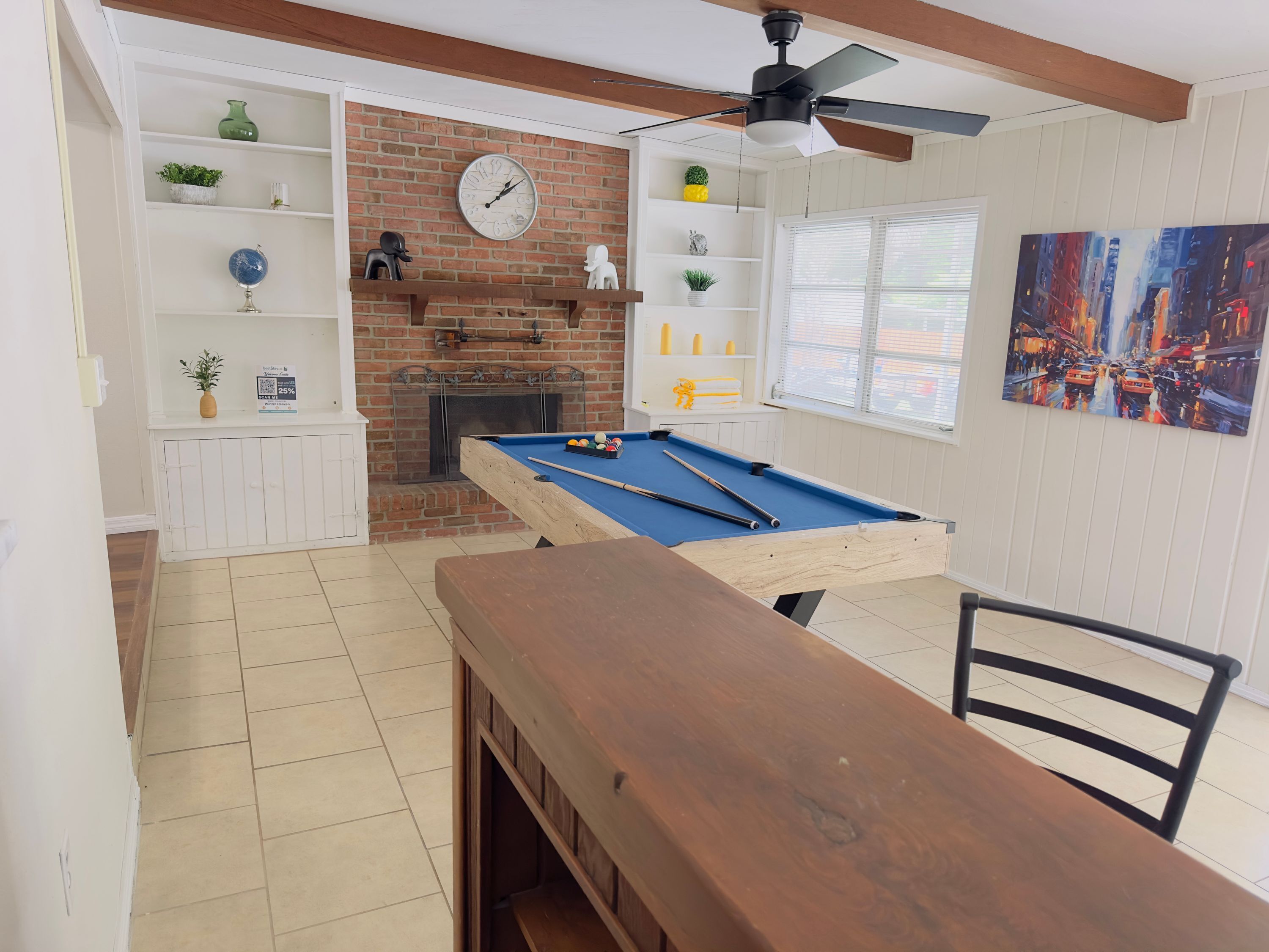 A pool table in a room with a brick fireplace, shelves with decorative items, and a painting on the wall.