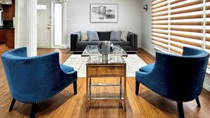 A living room with a dark grey sofa, two blue velvet armchairs, a glass coffee table, and a wooden floor. - The Hideaway Haven (Markham)