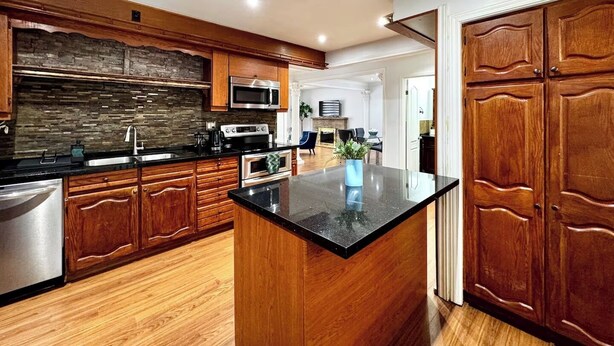 A modern kitchen with wooden cabinets, a granite countertop, stainless steel appliances, and a view into the living area. - The Hideaway Haven (Markham)