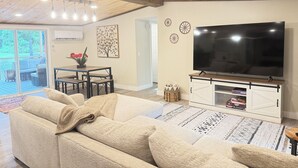 Een woonkamer met een grote bank, een televisie op een houten kast, een eettafel met stoelen en een plafond met houten balken en inbouwverlichting.