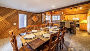 On aperçoit une salle à manger avec une table en bois dressée pour six personnes, entourée de chaises en bois. Au fond, on distingue une cuisine avec des placards en bois et un four à micro-ondes.
