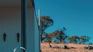 Un edificio bianco con una porta in metallo e un lavandino. Si vedono alberi e un paesaggio arido sullo sfondo.