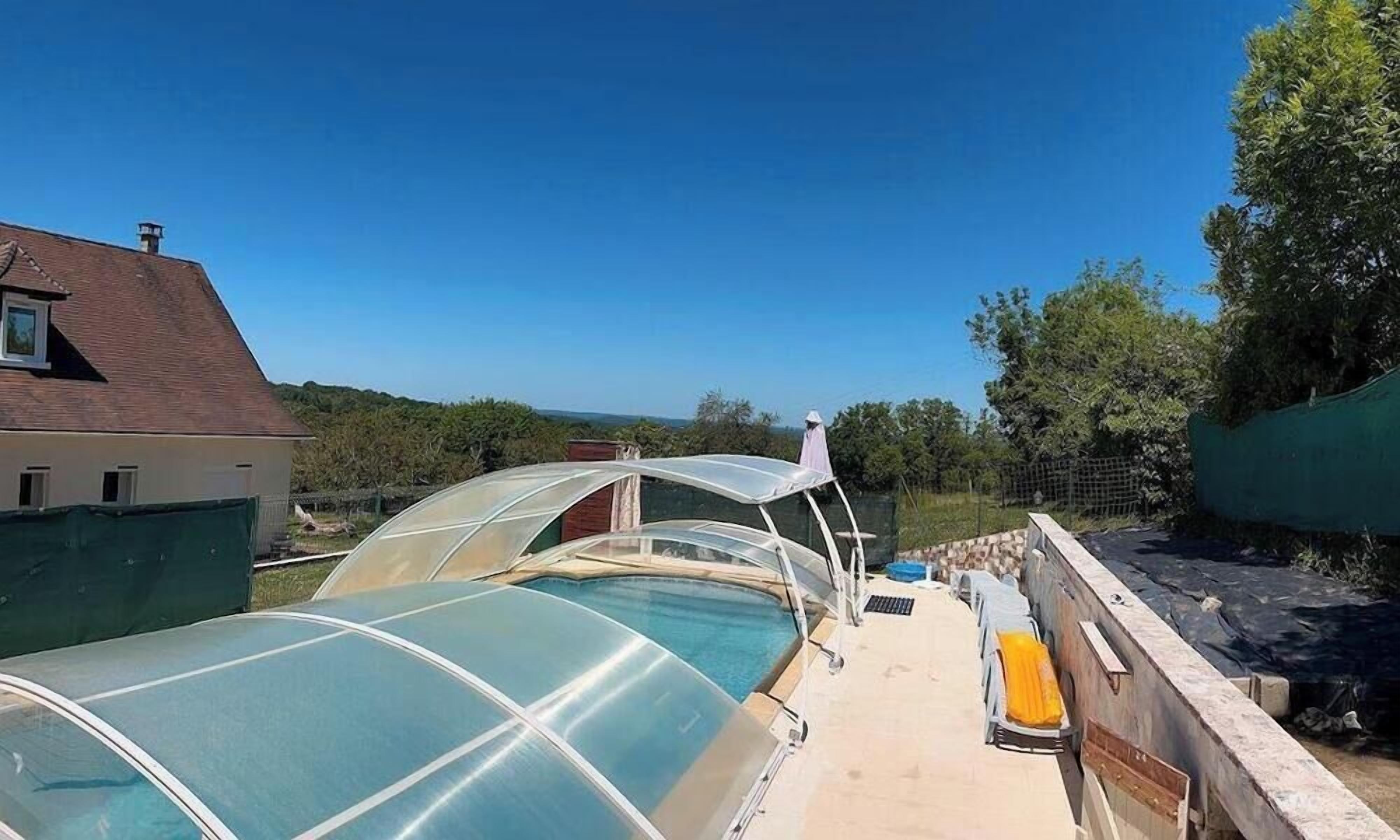 Une terrasse sur le toit avec une piscine, protégée par de grandes structures transparentes, entourée d’arbres et d’une maison.