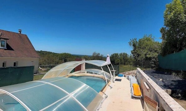 Una azotea con piscina, protegida por estructuras transparentes, rodeada de árboles y una casa.