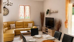 Ein Essbereich mit Tisch für vier Personen, einer Couch, einem Fernseher und einem Fenster mit Vorhängen.