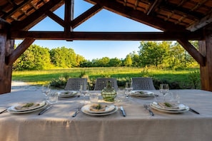 An outdoor dining area with a table set for four, surrounded by chairs, under a wooden structure with a view of a green field and trees.