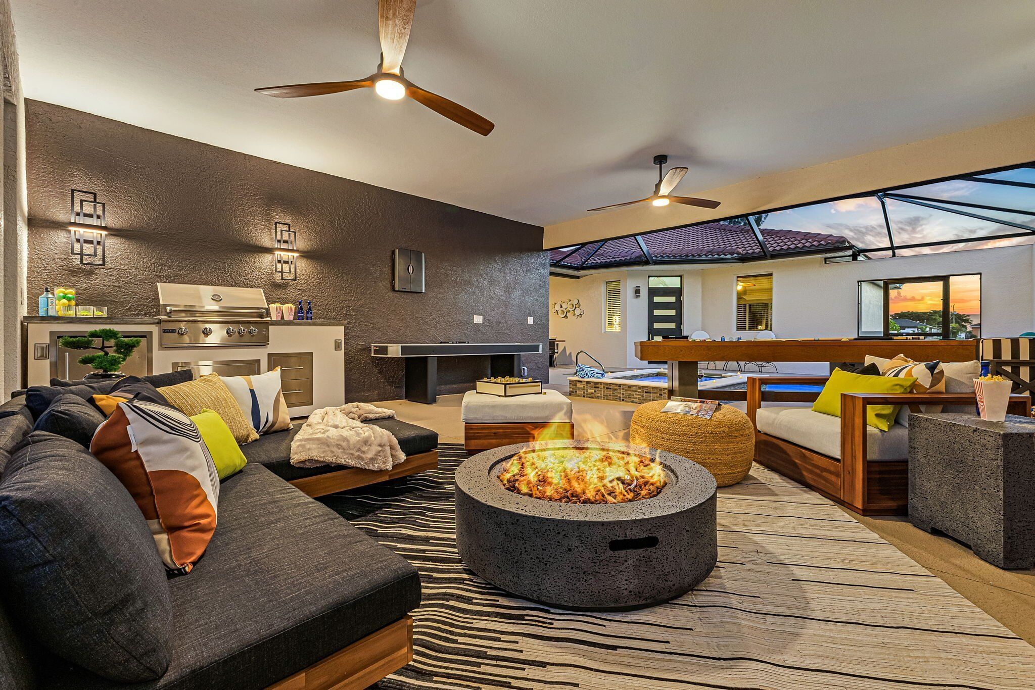 A modern living room with a stone fire pit, a kitchen area, and a view of the sunset through large windows.