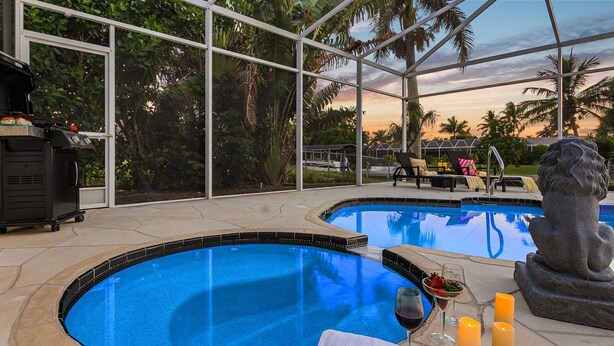 A pool area with a hot tub and a statue, surrounded by glass walls and palm trees. - Heated Pool & Spa with Dock, Tiki Hut & Gulf Access - Coral Reef - Roelens (Cape Coral)