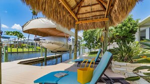 A poolside area with lounge chairs, a thatched roof, and a water feature. - Heated Pool & Spa with Dock, Tiki Hut & Gulf Access - Coral Reef - Roelens (Cape Coral)