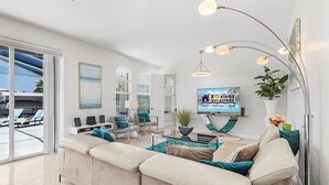 A modern living room with a beige sofa, a coffee table, and a TV. There is a large window with blinds, a plant, and a sign on the wall. - Heated Pool & Spa with Dock, Tiki Hut & Gulf Access - Coral Reef - Roelens (Cape Coral)