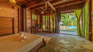 Eine Holzhütte mit Steinboden, einem Bett mit Handtuch, einem Stuhl und Blick auf einen Pool und Grünfläche.