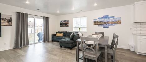 A spacious living area with a dark-colored sofa, a dining table with chairs, and a kitchen area with white cabinets.