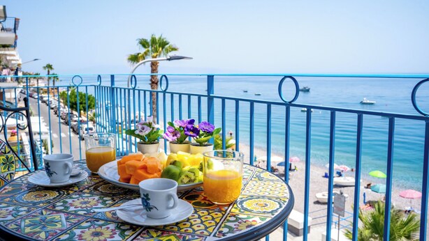Una mesa con alimentos y bebidas para el desayuno en un balcón con vista al mar.