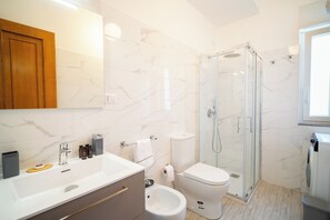 Une salle de bain moderne avec un mur en marbre, une douche en verre, un lavabo blanc et un bidet.