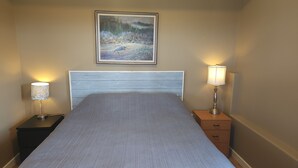 Un lit bien fait, recouvert d’une couvre-lit à rayures, deux lampes de chevet, un tableau encadré au-dessus de la tête de lit et une petite table de chevet en bois avec un tiroir.
