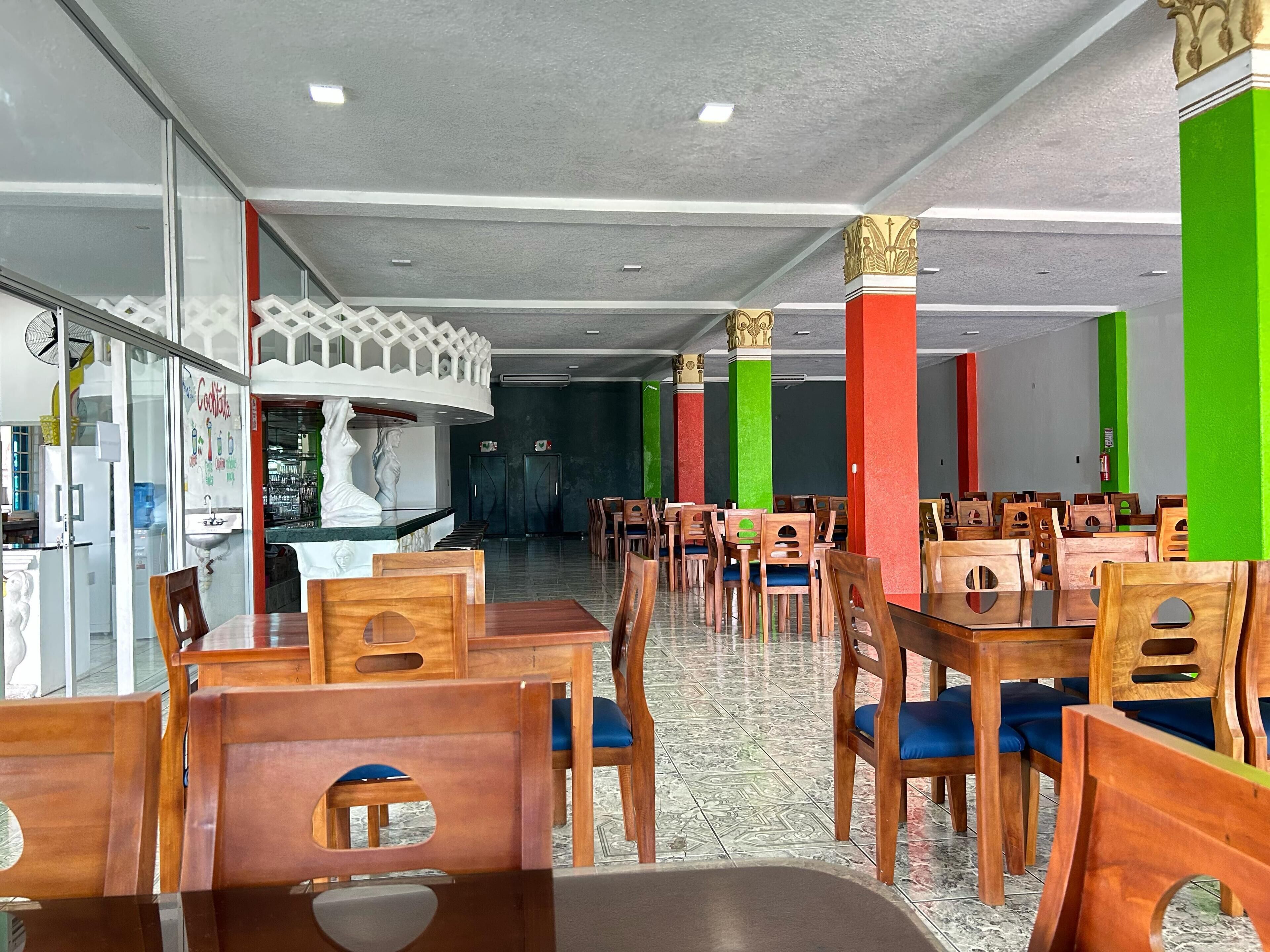 Un restaurant avec des chaises et des tables en bois, un comptoir orné d’une statue blanche et des piliers aux couleurs vives.