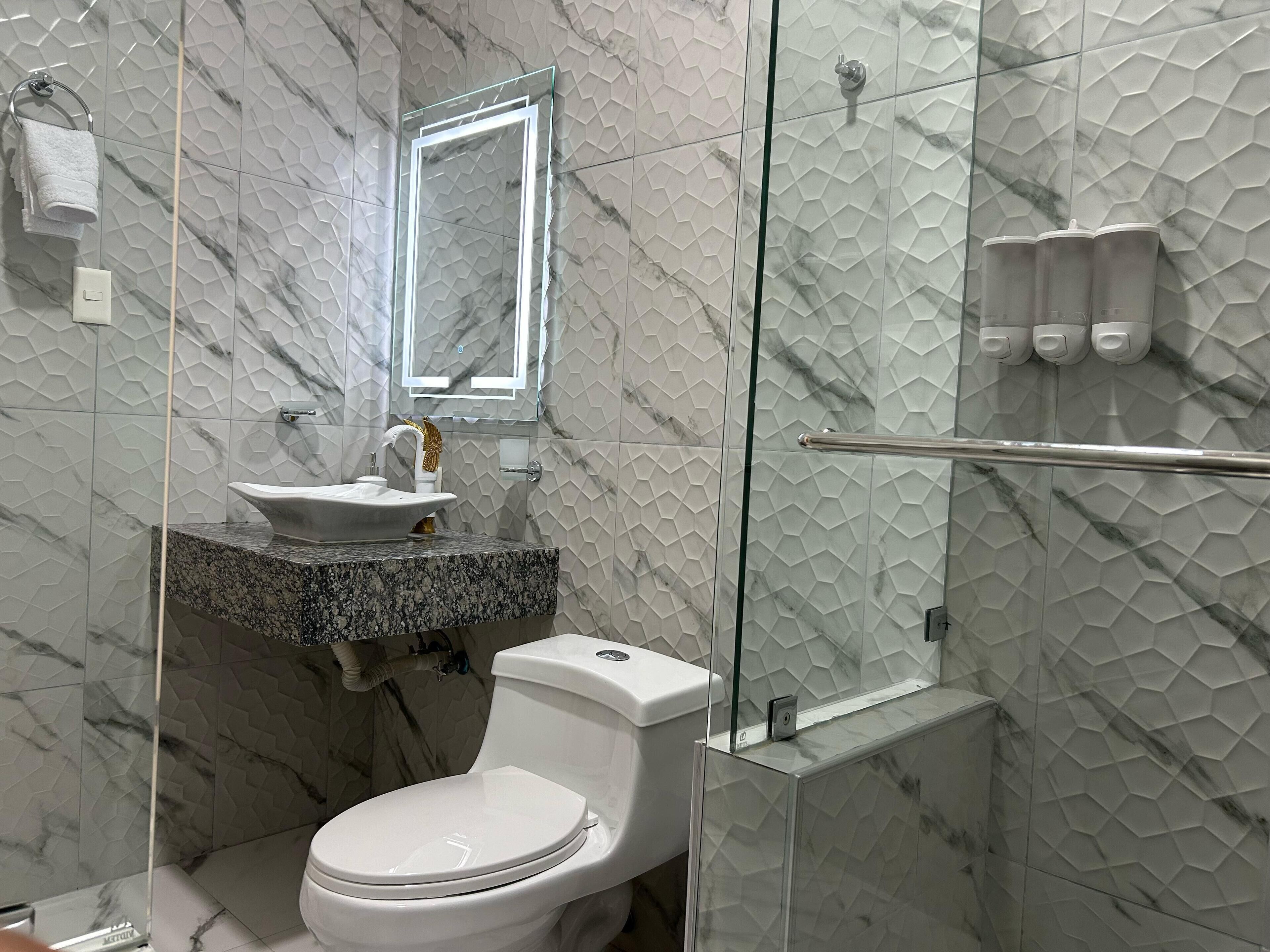 Ein Badezimmer mit Marmor-Musterwand, einem weißen Waschbecken mit Goldarmatur, einer Toilette und einer Duschkabine aus Glas.