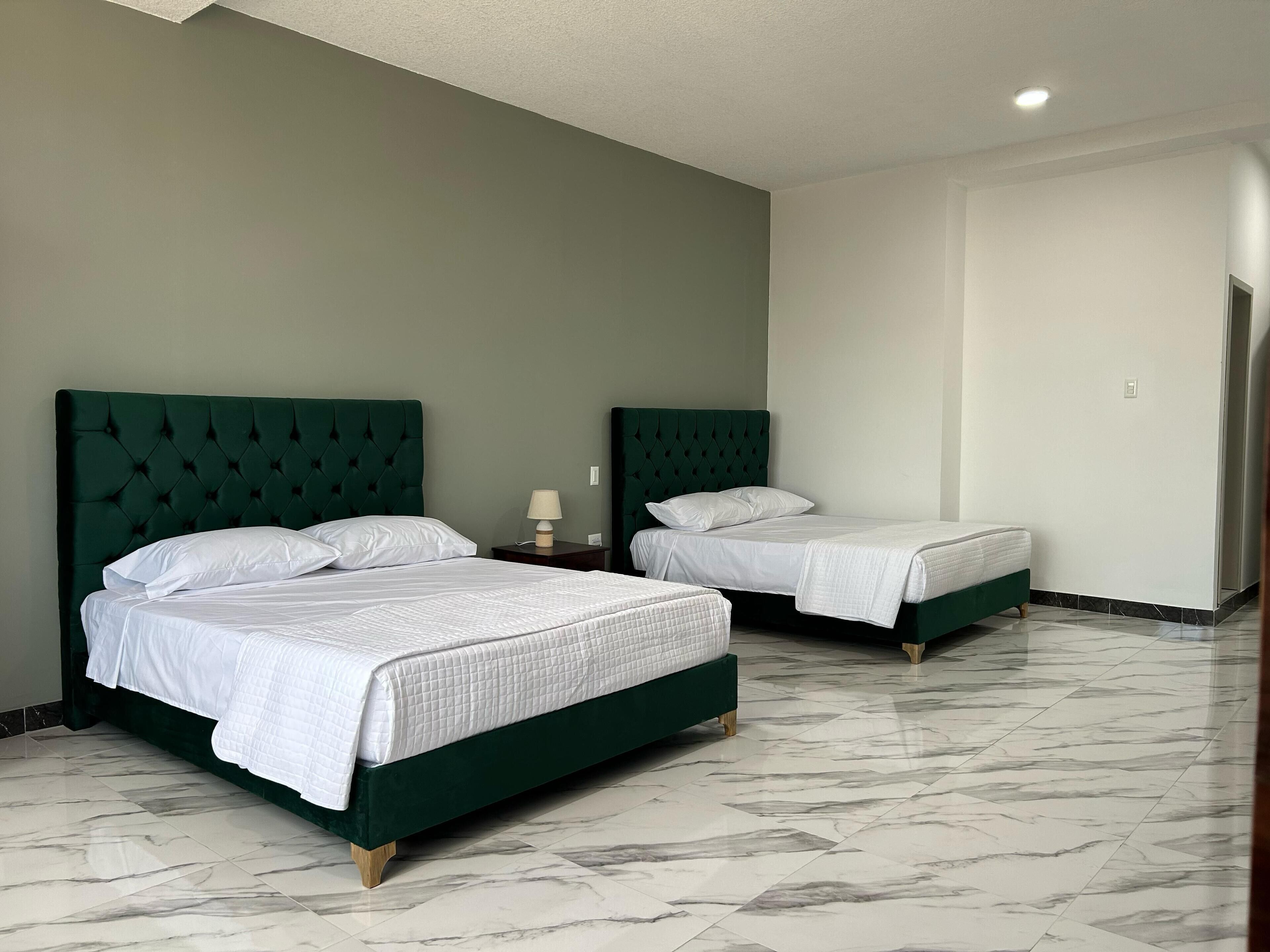 A hotel room with two beds, each with a green upholstered headboard and white bedding. The room has a marble-tiled floor and a plain white wall.