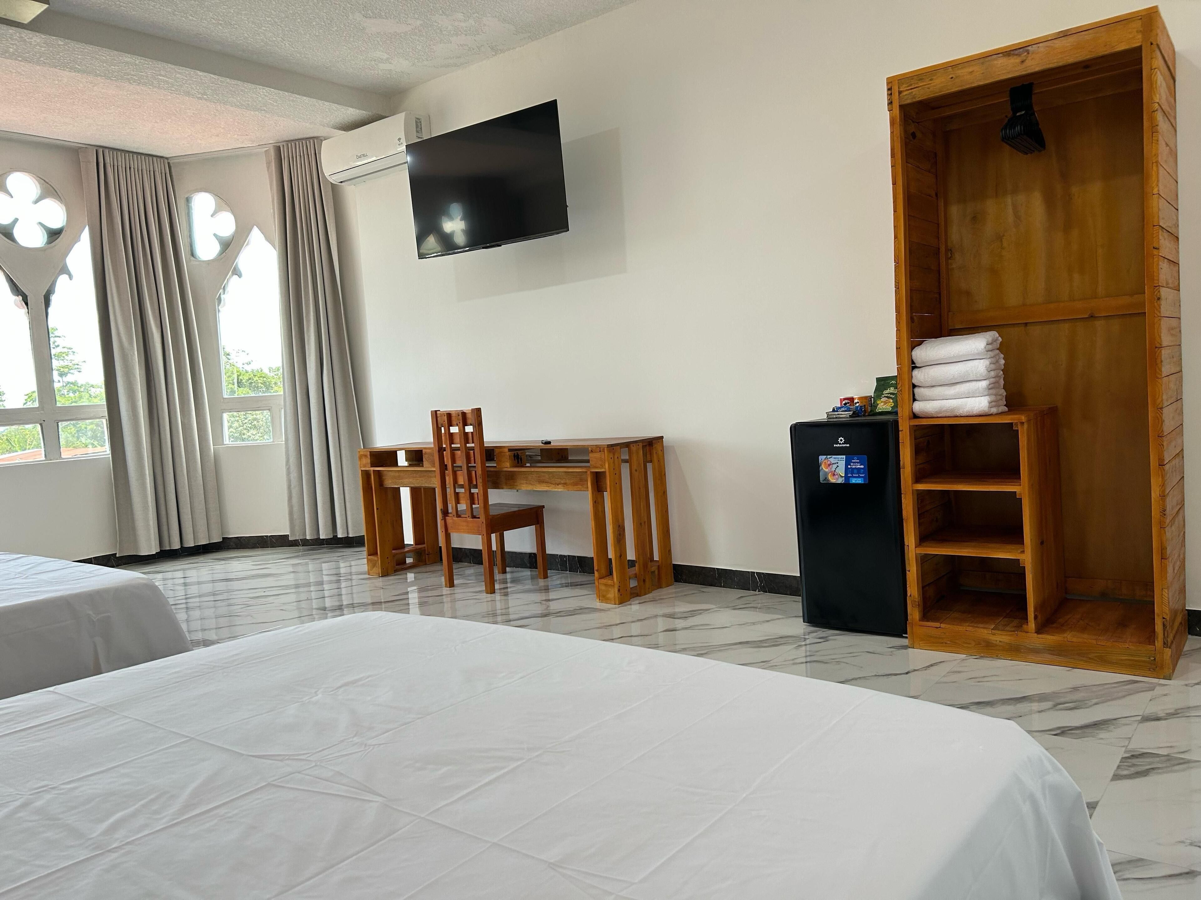 A hotel room with a bed, a desk, a chair, a TV, a wardrobe, and a small refrigerator.