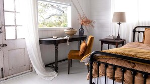 Un dormitorio con una cama, una silla, una mesa con un florero y una ventana con vistas.