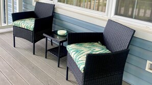 Een overdekt terras met twee rieten stoelen en een kleine tafel, met uitzicht op een woonwijk door grote ramen.