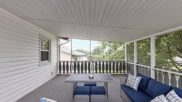 A covered porch with a table and seating area, featuring a railing and a view of the surrounding trees.