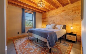 Une chambre avec un plafond en bois, un lit recouvert d’une couverture bleue, une table de chevet avec une lampe et un tapis à motifs.