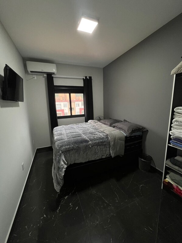 A bedroom with a bed, a TV, an air conditioner, a window with curtains, a fan, and a storage unit.