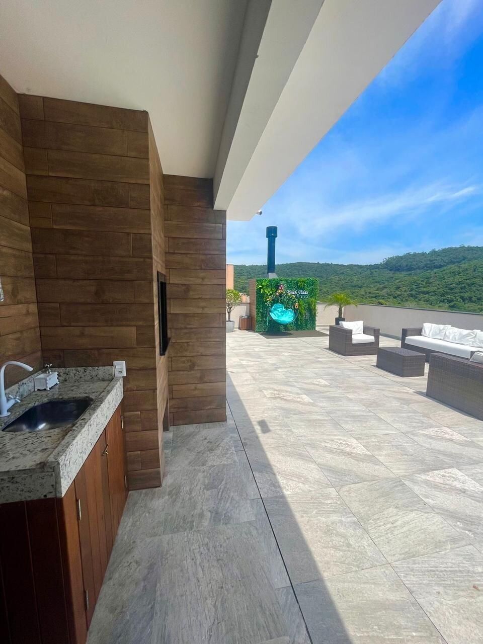 Una cocina moderna con un lavamanos integrado y armarios de madera, que da a una terraza con mobiliario exterior y vistas a una ladera verde y exuberante.