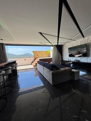 A modern living room with a large sofa, a flat-screen TV mounted on the wall, and a view of the outdoors.